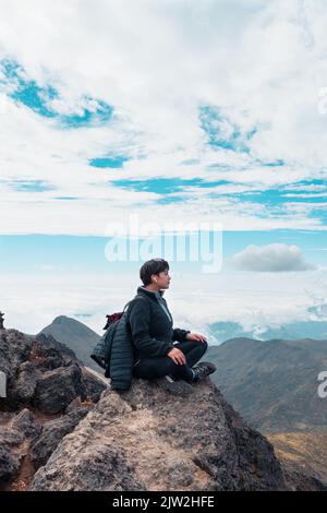 Vista laterale dell'escursionista femminile negli auricolari seduti guardando lontano sulla roccia a Padmasana, nelle Highlands, e ascoltando la meditazione mentre si rilassa e si fa yoga Foto Stock