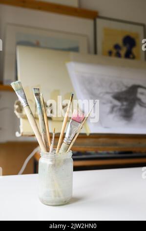 Vasetto di vetro sporco con set di pennelli posizionati su cavalletto di legno contro carta bianca in officina Foto Stock