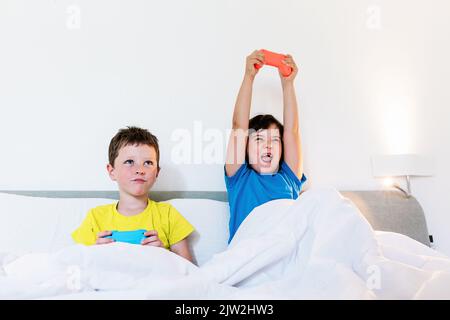Ragazza alzando le braccia con game pad in eccitazione mentre si gioca a video gioco con il fratello sconvolto nel letto in camera da letto luce Foto Stock