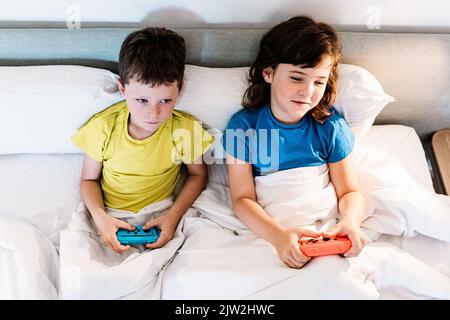 Da sopra i fratelli concentrati con gamepad che giocano il video gioco interessante mentre si siedono insieme nel letto comodo sotto coperta nella camera da letto a casa Foto Stock