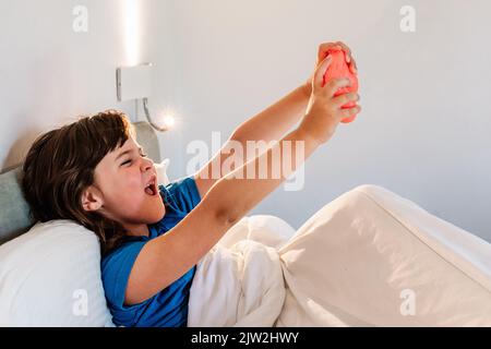 Vista laterale della ragazza entusiasta che alza le braccia con il game pad in eccitazione mentre si gioca a videogiochi a letto in camera da letto leggera Foto Stock