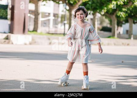 Corpo pieno sorridente ragazza carina in estate vestito a cavallo pattini a rotelle e guardare lontano mentre si passa la giornata estiva di sole sulla strada della città nel fine settimana Foto Stock