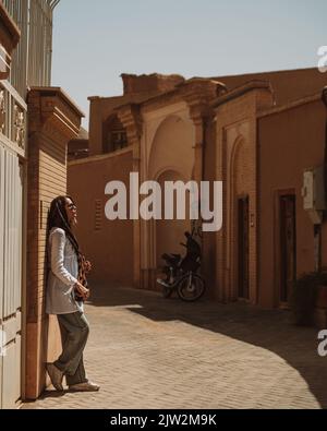 Vista laterale del corpo intero di una donna viaggiatore in un velo in piedi vicino ai tipici edifici shabby nella città dell'Iran il giorno d'estate Foto Stock