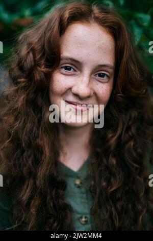 Giovane donna fidata e sicura con lunghi capelli ricci rossi in abiti casual in piedi vicino all'arbusto sorridendo alla macchina fotografica Foto Stock