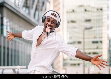 Felice uomo afroamericano in camicia bianca e berretto che sparge le braccia e mostra la lingua mentre ascolta musica e danze sullo sfondo sfocato della città Foto Stock