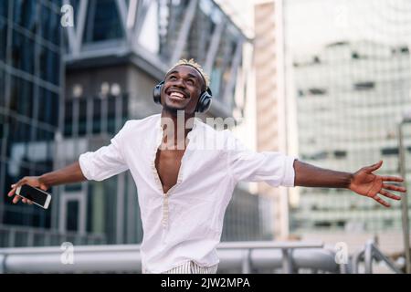 Felice uomo afroamericano in camicia bianca che sparge le braccia mentre ascolti musica e danze su sfondo sfocato della strada cittadina di giorno Foto Stock