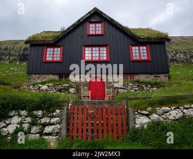 Da sotto cottage nero con finestre rosse e porta situata su collina erbosa dietro cancello di legno contro cielo nuvoloso in giorno grigio nel villaggio sull'isola di Faroe Foto Stock