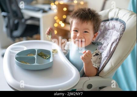 Bambino che mangia da solo nella sua seggiolone a casa. Adorabile bambino di un anno che ha un pasto tenendo il cibo nelle sue mani e mangiare a casa Foto Stock