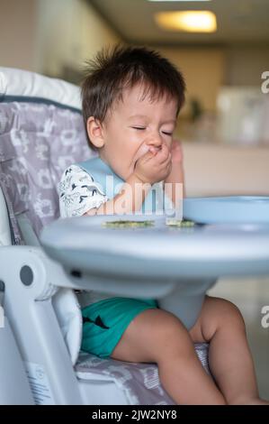 Bambino che mangia da solo nella sua seggiolone a casa. Adorabile bambino di un anno che ha un pasto tenendo il cibo nelle sue mani e mangiare a casa Foto Stock