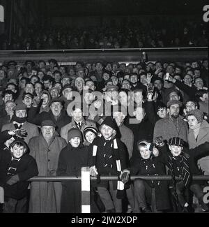 1960s, storici, tifosi di calcio in piedi insieme su una terrazza tradizionale per guardare una partita, in una partita serale... cappotti, cappellini piatti per gli uomini, cappotti duffel e cappelli bobble per i giovani, l'era prima di replica camicie! Foto Stock