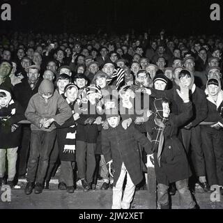 1960s, storico, tifosi di calcio, incontro serale, cappotti, cappellini piatti per gli uomini, cappotti e cappelli bobly per i più piccoli, non una replica camicia in vista! Foto Stock