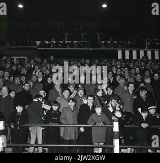 1960s, storici, tifosi di calcio in piedi insieme su una terrazza tradizionale per guardare una partita, in una partita serale... cappotti, cappellini piatti per gli uomini, cappotti duffel e cappelli bobble per i giovani, l'era prima di replica camicie! Foto Stock