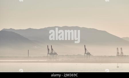 Tramonto nel porto di Malaga con nebbia causata da umidità con gru, navi a vela e montagne dietro Foto Stock