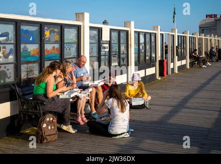 Un gruppo di famiglie si siede per mangiare pesce tradizionale e patatine fritte al mare sul Molo di Worthing, West Sussex, Regno Unito. Foto Stock