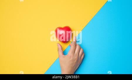 Il cuore rosso e la mano di una donna lo raggiunge su uno sfondo blu-giallo Foto Stock