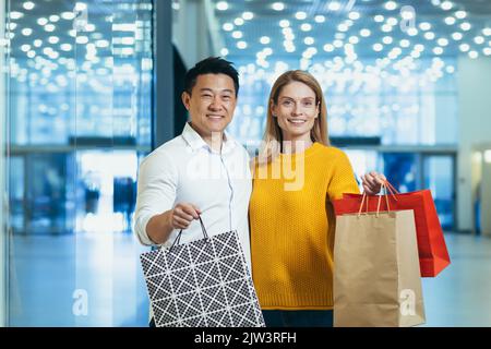 Giovane famiglia diversa uomo asiatico e donna bionda shopping in supermercato, ritratto di coppia acquirenti guardando la macchina fotografica e sorridendo e tenendo borse colorate con merci e shopping. Foto Stock