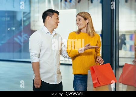 coppia multirazziale uomo asiatico e donna caucasica che camminano insieme nel centro commerciale dopo aver fatto shopping con borse colorate in mano. passeggiando davanti alle vetrine del negozio con vestiti famiglia allegra che abbraccia il sorriso. Foto Stock