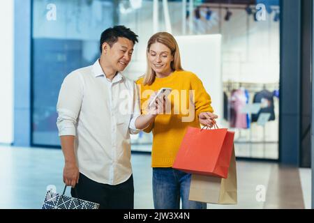 Coppia asiatica e donna bionda diversi in supermercato vestiti, guardando lo schermo del telefono cellulare, scegliendo shopping, tenendo borse e regali colorati. Foto Stock
