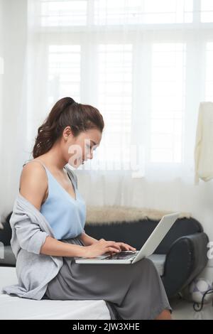 Bella giovane donna vietnamita seduta sul bordo del letto e al lavoro sul laptop Foto Stock