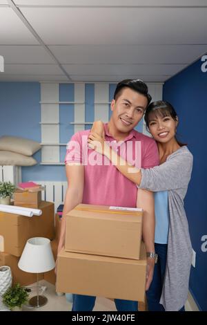 Ritratto di giovane coppia asiatica in piedi in nuovo piano, tenendo scatole di cartone con roba di casa e sorridendo alla macchina fotografica con gioia Foto Stock