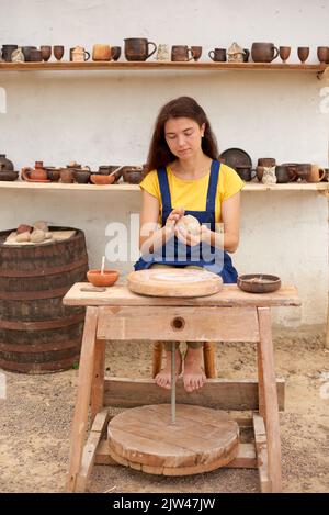 Donna in grembiule blu e T-shirt gialla produce prodotti in argilla su ruote meccaniche in vasaio Foto Stock