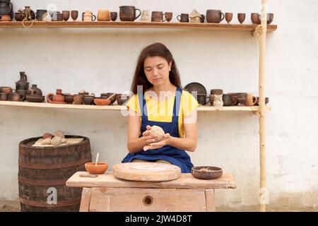 Donna in grembiule blu e T-shirt gialla lavora sulla ruota meccanica del vasaio sullo sfondo di scaffali di prodotti in argilla Foto Stock