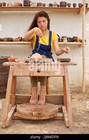 Donna in grembiule blu e T-shirt gialla lavora sulla ruota meccanica del vasaio sullo sfondo di scaffali di prodotti in argilla Foto Stock