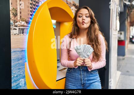 Giovane donna ispanica che prende denaro da atm facendo pesce faccia con la bocca e squinting occhi, pazzo e comico. Foto Stock