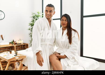 Giovane coppia latina che indossa un asciugamano in piedi al centro di bellezza sorridendo guardando al lato e fissando via pensando. Foto Stock