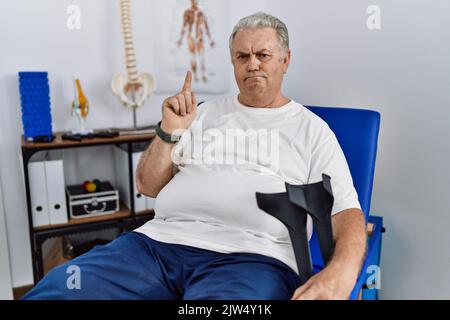 L'uomo caucasico anziano alla clinica di fisioterapia che tiene le stampelle che indicano in su osservare triste e sconvolto, indicando la direzione con le dita, infelice e depres Foto Stock