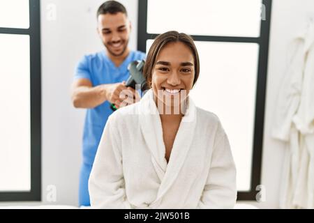 Latino uomo e donna che indossano la fisioterapia uniforme massaggiante collo con pistola a percussione al centro di bellezza Foto Stock
