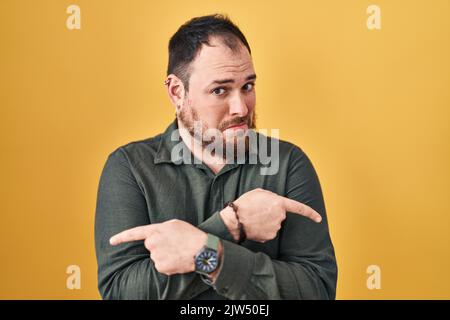 Più misura uomo ispanico con barba in piedi su sfondo giallo che punta ad entrambi i lati con le dita, diverso senso non sono d'accordo Foto Stock