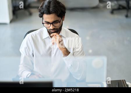 Uomo d'affari indiano serio occupato che analizza i dati di mercato, guardando il computer. Foto Stock