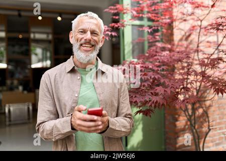 Sorridente uomo anziano di mezza età utilizzando un telefono in piedi all'aperto. Foto Stock