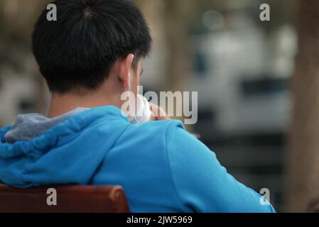 L'uomo asiatico con una felpa con cappuccio blu ascolta la musica con gli auricolari mentre beve una tazza di caffè fuori da un bar nel bel mezzo della giornata. Foto dal retro. Foto Stock