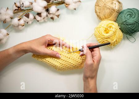 Le mani della donna sono vestiti del bambino di maglia da filato giallo, su un tavolo bianco, vista dall'alto. Foto Stock