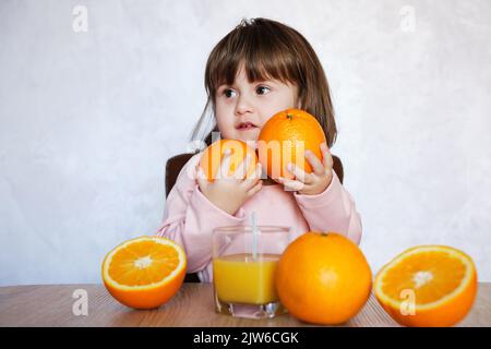 Ritratto di una bambina gioca con un arance su un tavolo. Ragazza piccola felice con un bicchiere di succo. Stile di vita sano. Foto Stock