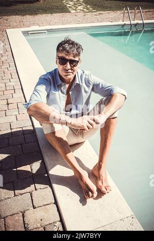 Il giovane indossa occhiali da sole, una camicia bianca e pantaloni beige e si siede vicino alla piscina. Bell'uomo d'affari prende un momento di relax presso il Foto Stock