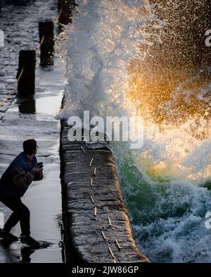 IN ARRIVO! Grande onda d'acqua che sbatte nel muro del porto a Porthleven appena mancante un uomo sul suo telefono. Preso al tramonto. Foto Stock