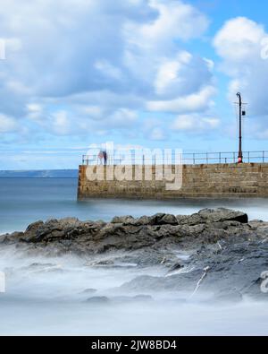 La velocità dell'otturatore è ridotta per catturare le onde morbide che si infrangono sul molo di Porthleven, mentre i pescatori e i visitatori esplorano il molo. Foto Stock