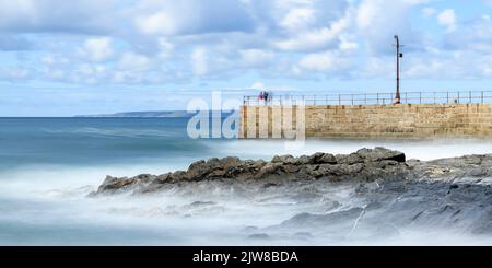 La velocità dell'otturatore è ridotta per catturare le onde morbide che si infrangono sul molo di Porthleven, mentre i pescatori e i visitatori esplorano il molo. Foto Stock