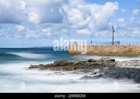 La velocità dell'otturatore è ridotta per catturare le onde morbide che si infrangono sul molo di Porthleven, mentre i pescatori e i visitatori esplorano il molo. Foto Stock