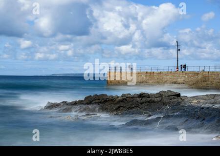 La velocità dell'otturatore è ridotta per catturare le onde morbide che si infrangono sul molo di Porthleven, mentre i pescatori e i visitatori esplorano il molo. Foto Stock