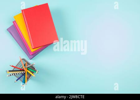 Libri colorati e matite colorate in maglia metallica su sfondo azzurro. Vista dall'alto. Istruzione, apprendimento delle lingue, concetto di ritorno a scuola Foto Stock