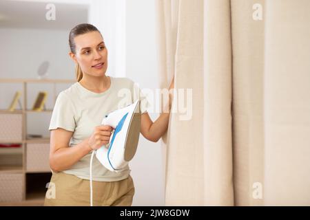 Giovane donna che stira le tende a casa Foto Stock