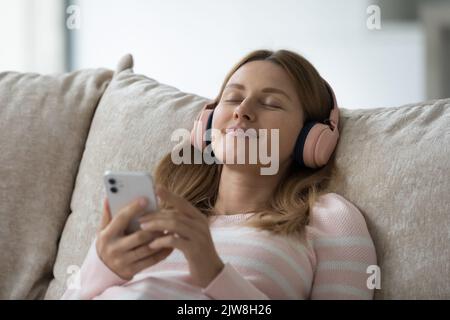 Donna utente di gadget millennial rilassato e spensierato che indossa cuffie grandi Foto Stock