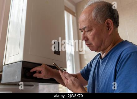 Un uomo anziano che abbaglia la caldaia dell'acqua calda per risparmiare denaro cercando di far fronte a un aumento drammatico dei costi energetici. Foto Stock