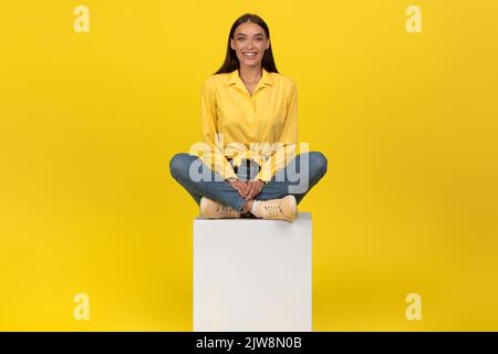 Felice giovane femmina seduto su cubo in posa su sfondo giallo Foto Stock