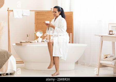 Donna mista che tiene in vaso crema idratante in bagno moderno Foto Stock