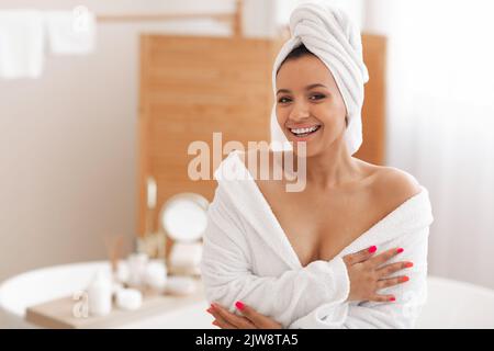 Attraente misto femmina sorridente alla fotocamera indossando accappatoio in bagno Foto Stock